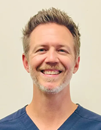 Smiling man headshot with short blond hair, light stubble, navy scrub top